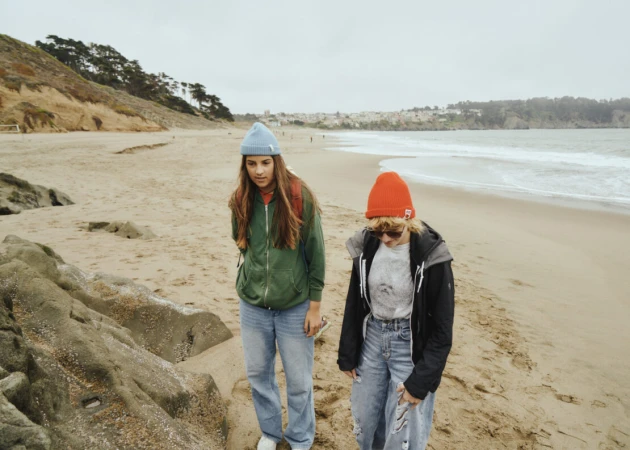 Two young women walking along a sandy beach under an overcast sky, wearing beanies and casual jackets.