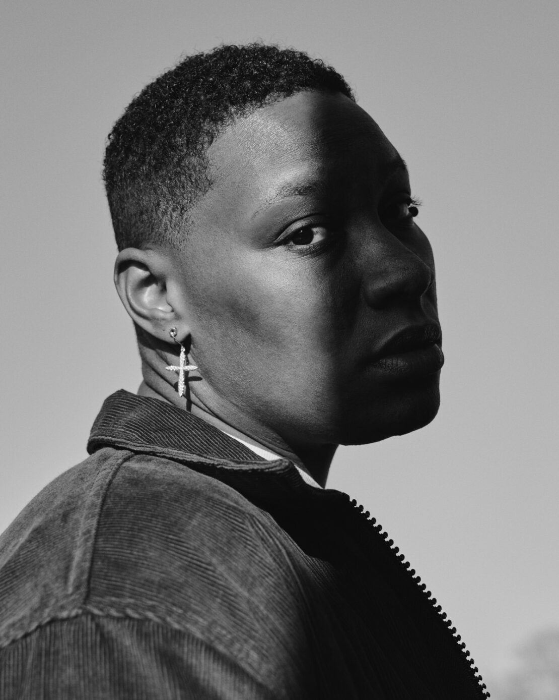Black and white close-up portrait of a stylish African American male wearing a corduroy jacket and a cross earring under bright sunlight.