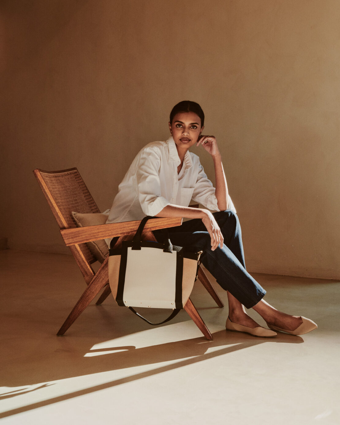 A woman in a white shirt sits in a wooden chair, a tote bag next to her, illuminated by soft, natural light in a minimalist indoor setting.