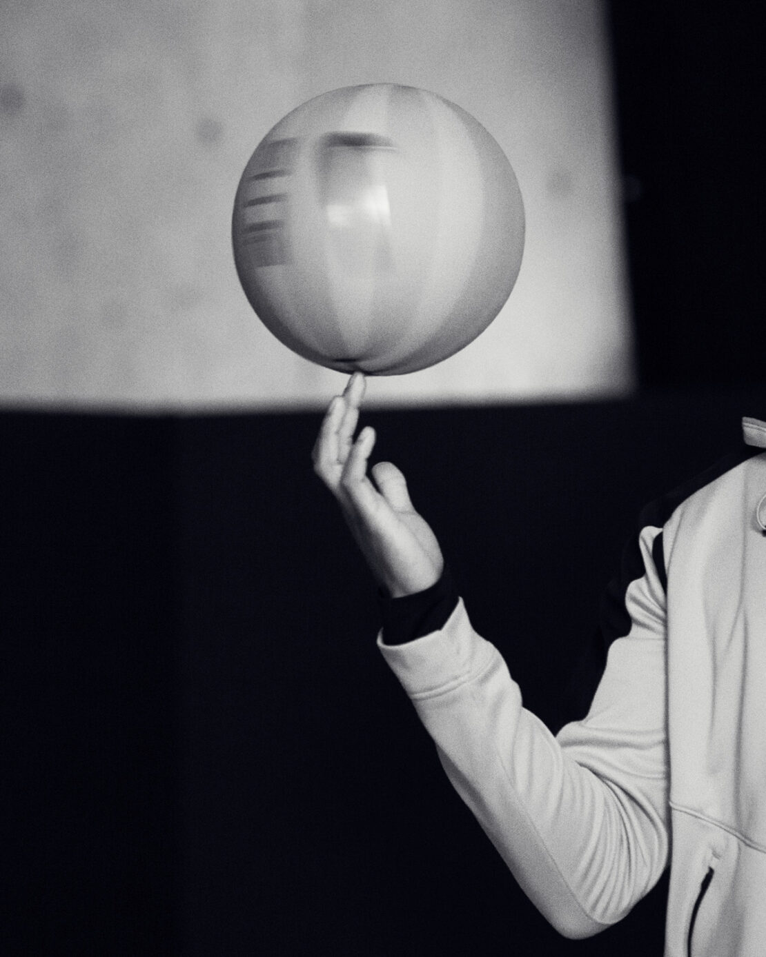 Monochrome photo of a spinning ball balanced on a person's fingertip with hand and arm visible, set against a dark background.