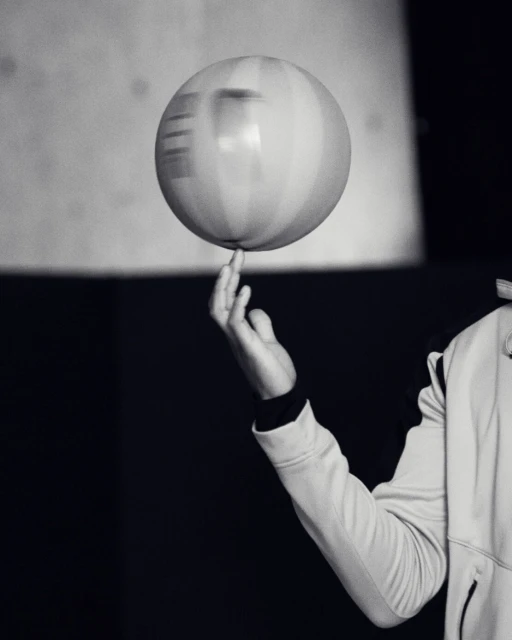 Monochrome photo of a spinning ball balanced on a person's fingertip with hand and arm visible, set against a dark background.