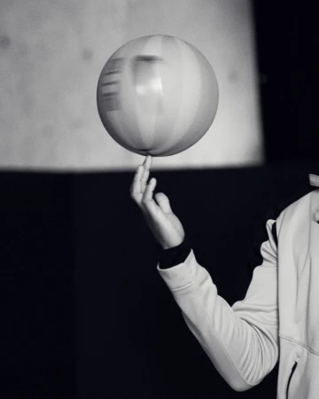 Monochrome photo of a spinning ball balanced on a person's fingertip with hand and arm visible, set against a dark background.