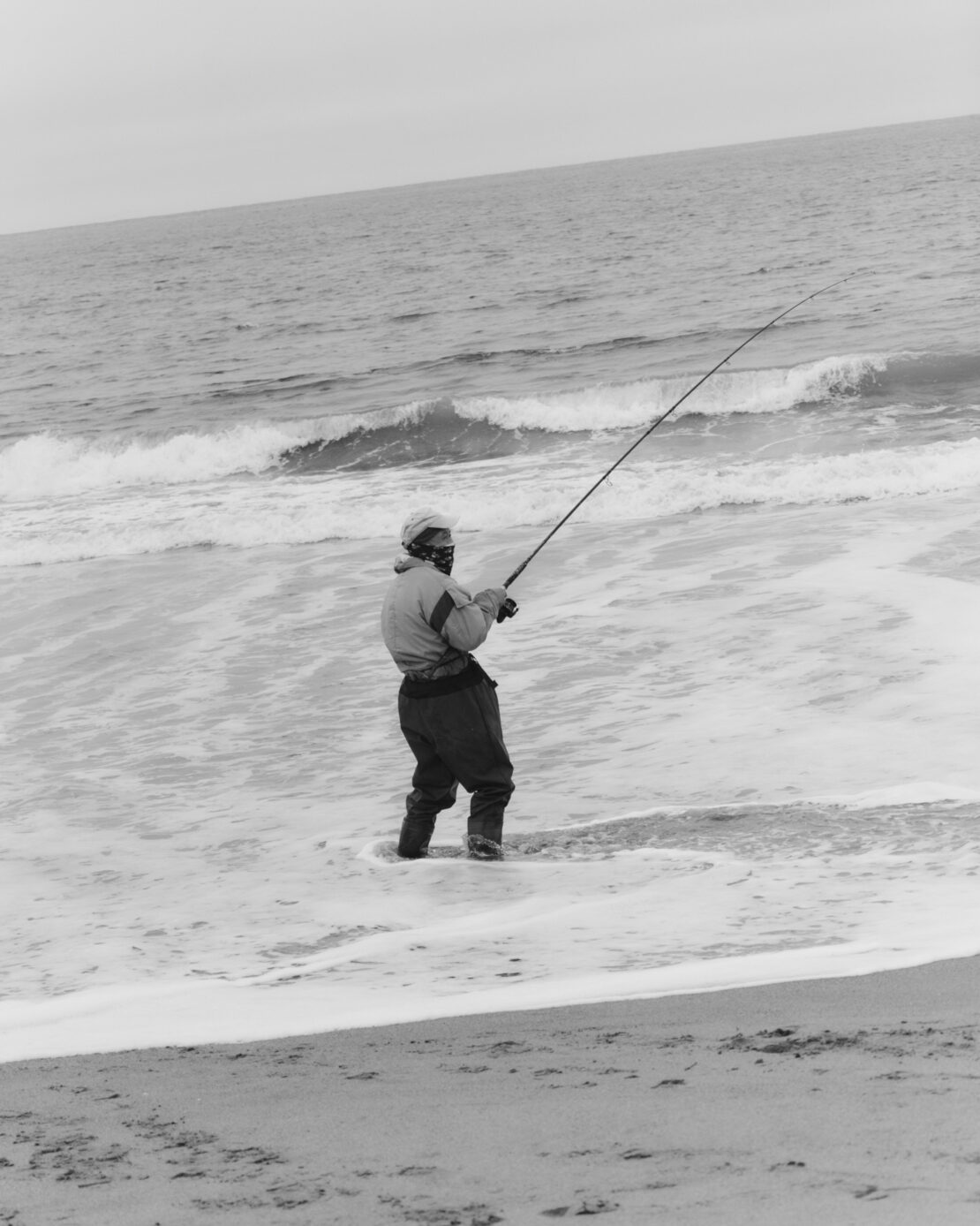 A person fishes while standing in the surf, casting a fishing rod out into the ocean under an overcast sky.