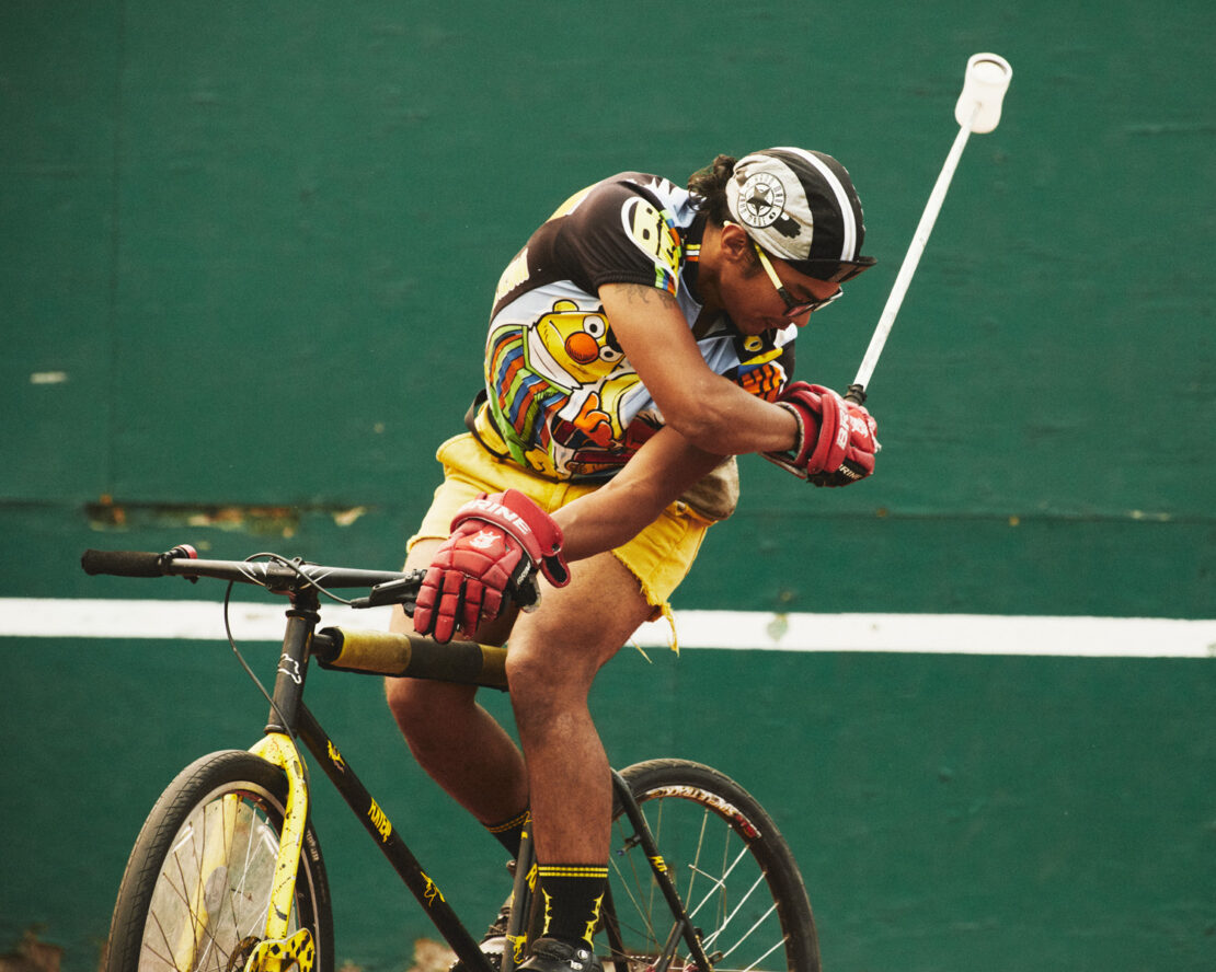 Adult male rides a bicycle outdoors against a green wall, wearing a printed shirt, yellow shorts, helmet, and gloves, holding a lacrosse stick.