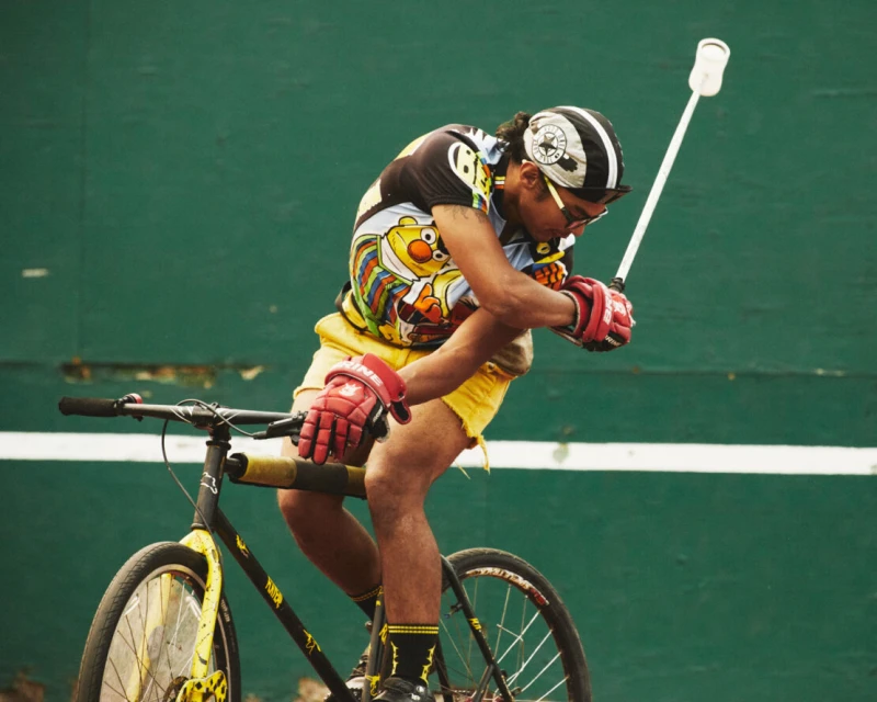 Adult male rides a bicycle outdoors against a green wall, wearing a printed shirt, yellow shorts, helmet, and gloves, holding a lacrosse stick.
