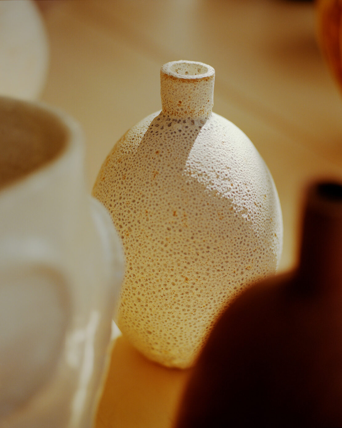 Still life shot of a collection of three ceramic vases in earth tones and white sitting on a flat surface in soft, diffused lighting.