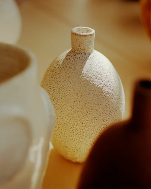Still life shot of a collection of three ceramic vases in earth tones and white sitting on a flat surface in soft, diffused lighting.
