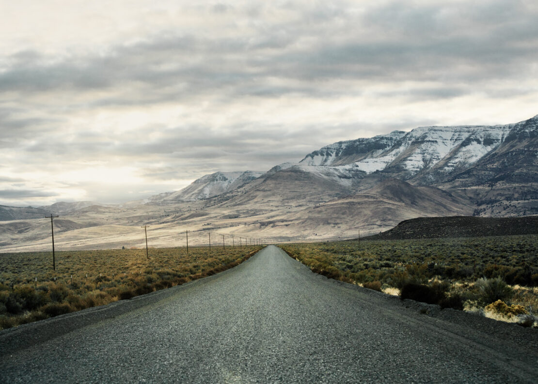 A desolate gravel road stretches into the distance towards mountains, beneath a cloudy sky; power lines line the roadside.