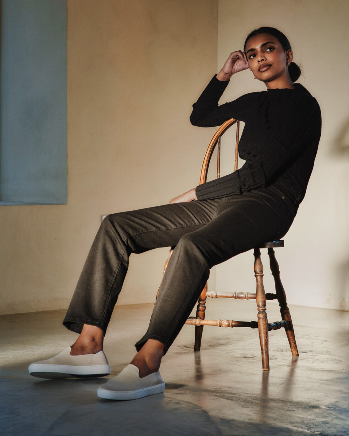 A brunette woman sits on a wooden chair in a studio, wearing a black top, dark pants, and beige sneakers. The background is a muted beige.