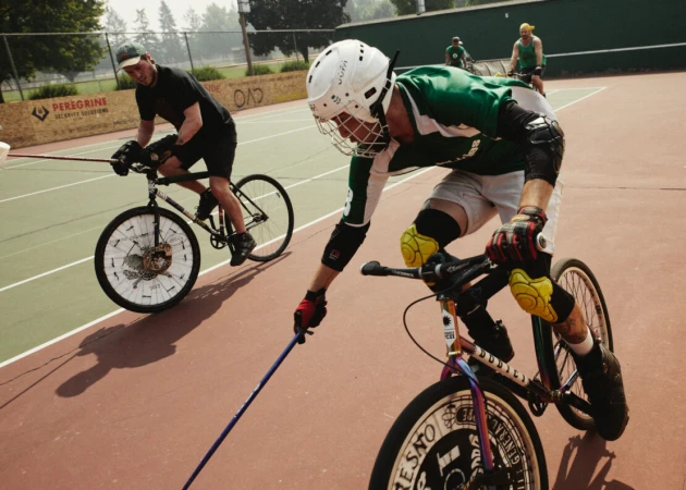 Two men on bicycles with helmets and protective gear compete on an outdoor court while holding mallets during a bike polo match.