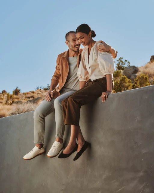 Outdoor lifestyle shot of a happy couple in neutral clothing sitting on a concrete wall with tan grass in the background under blue sky.