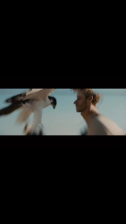 Osprey flies near a shirtless man outdoors, creating a captivating nature scene under clear sky. Motion blur adds a sense of dynamism.