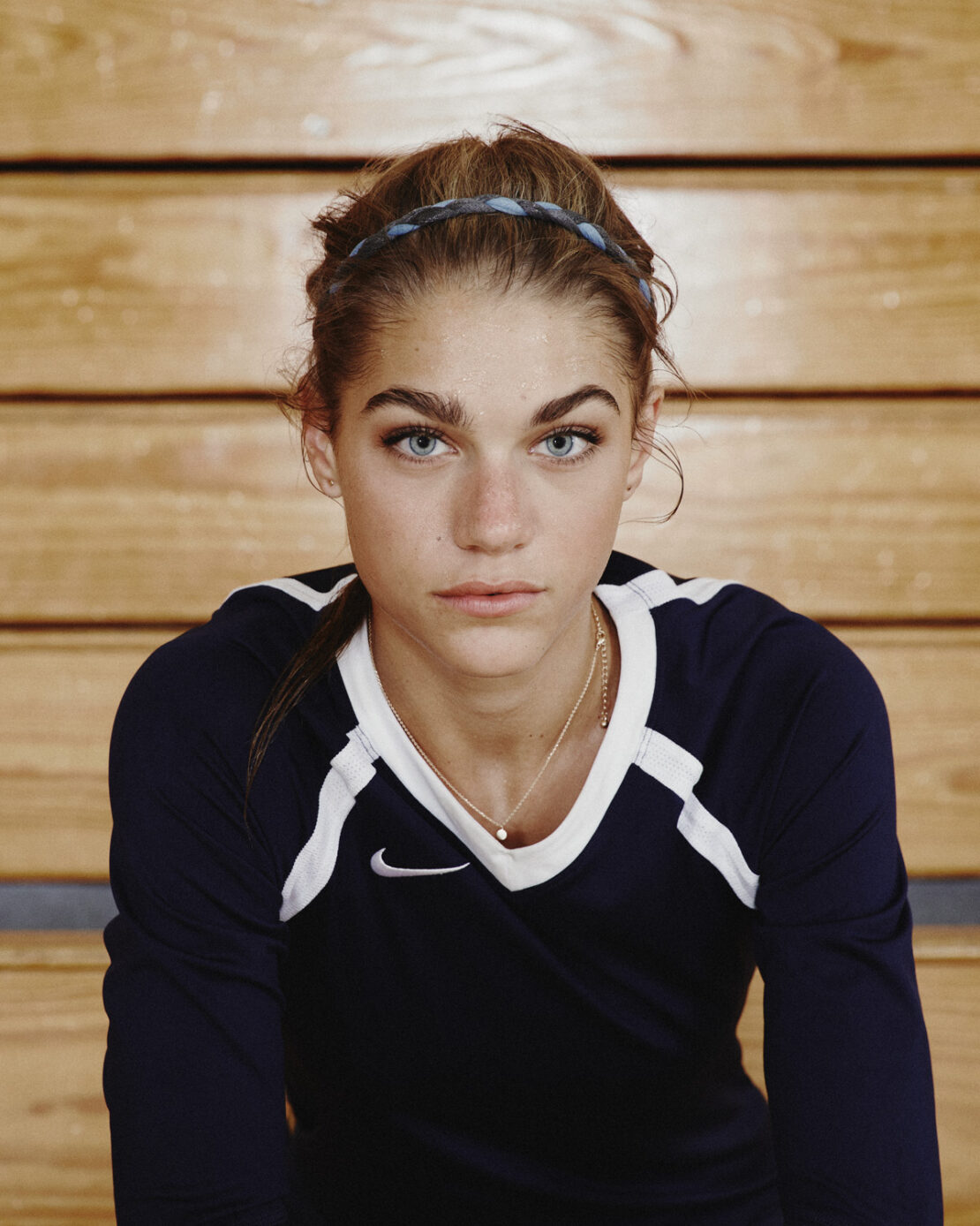 Portrait of a female athlete with a blue headband wearing a navy blue sports jersey against a wooden wall background.