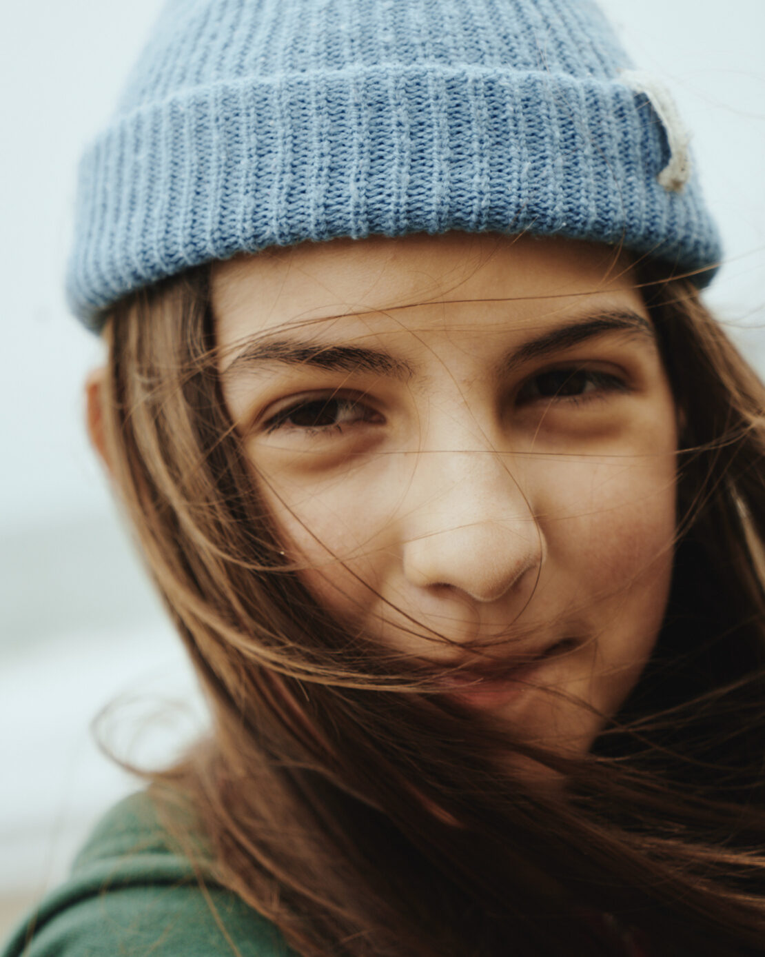 Close-up portrait of a young person wearing a blue ribbed beanie and green top with hair blowing across their face outdoors.