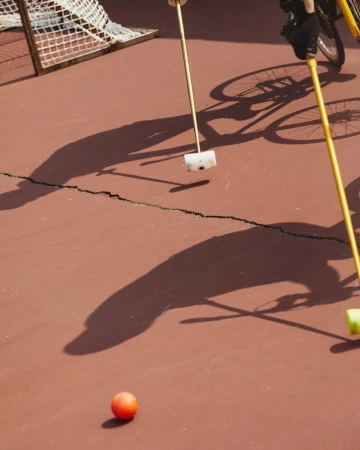 A bright orange ball sits on a reddish-brown court near a white and black net. People play bike polo with yellow mallets and bicycles.