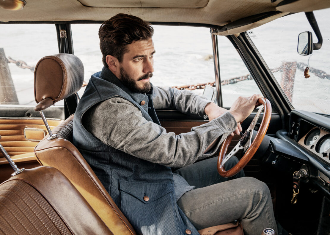 Man driving a vintage car, wearing a blue vest and gray shirt, with a focused expression, capturing a classic automotive and fashion moment.