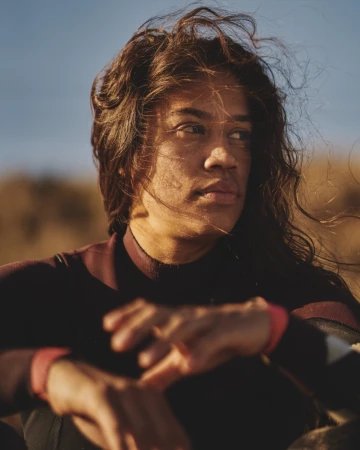 Portrait of a young adult with long brown hair wearing a wetsuit and sitting outdoors in warm sunlight, looking away from the camera.