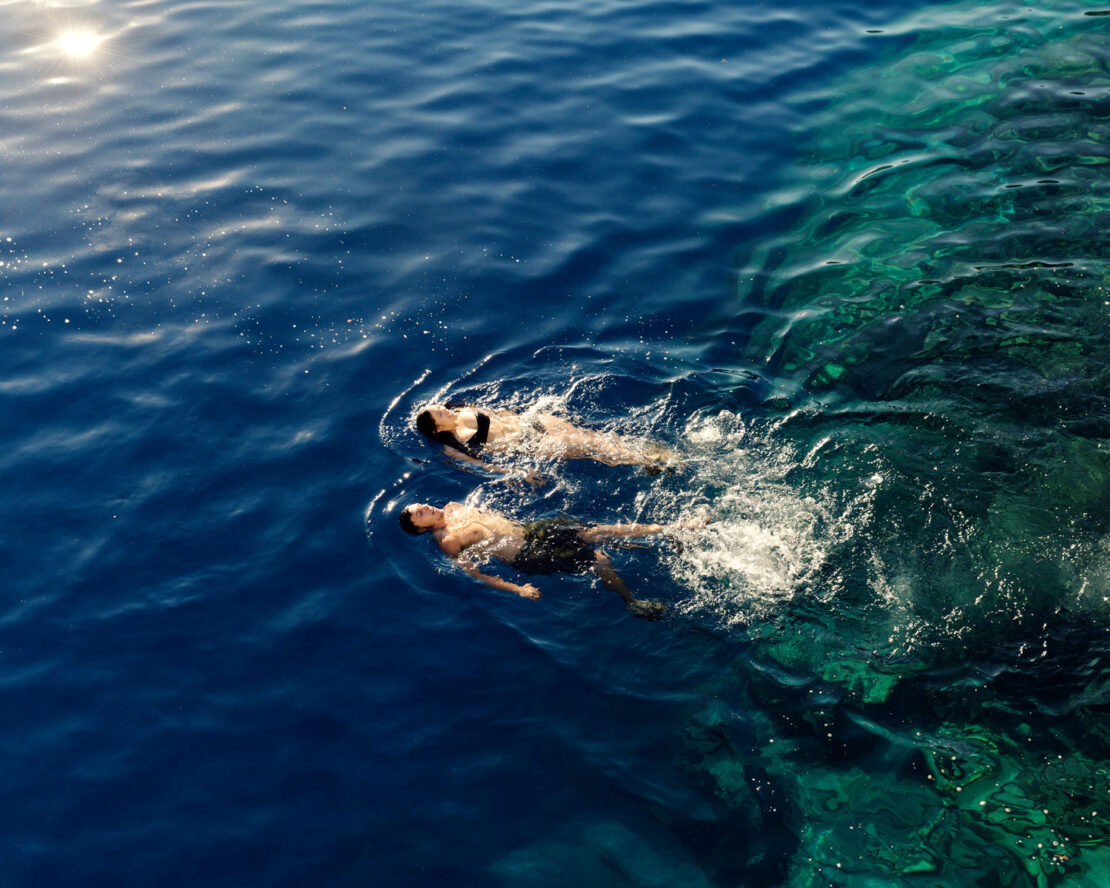 Aerial view of a man and woman swimming in deep blue ocean waters, sunlight reflecting on the surface, near a rocky coastline.