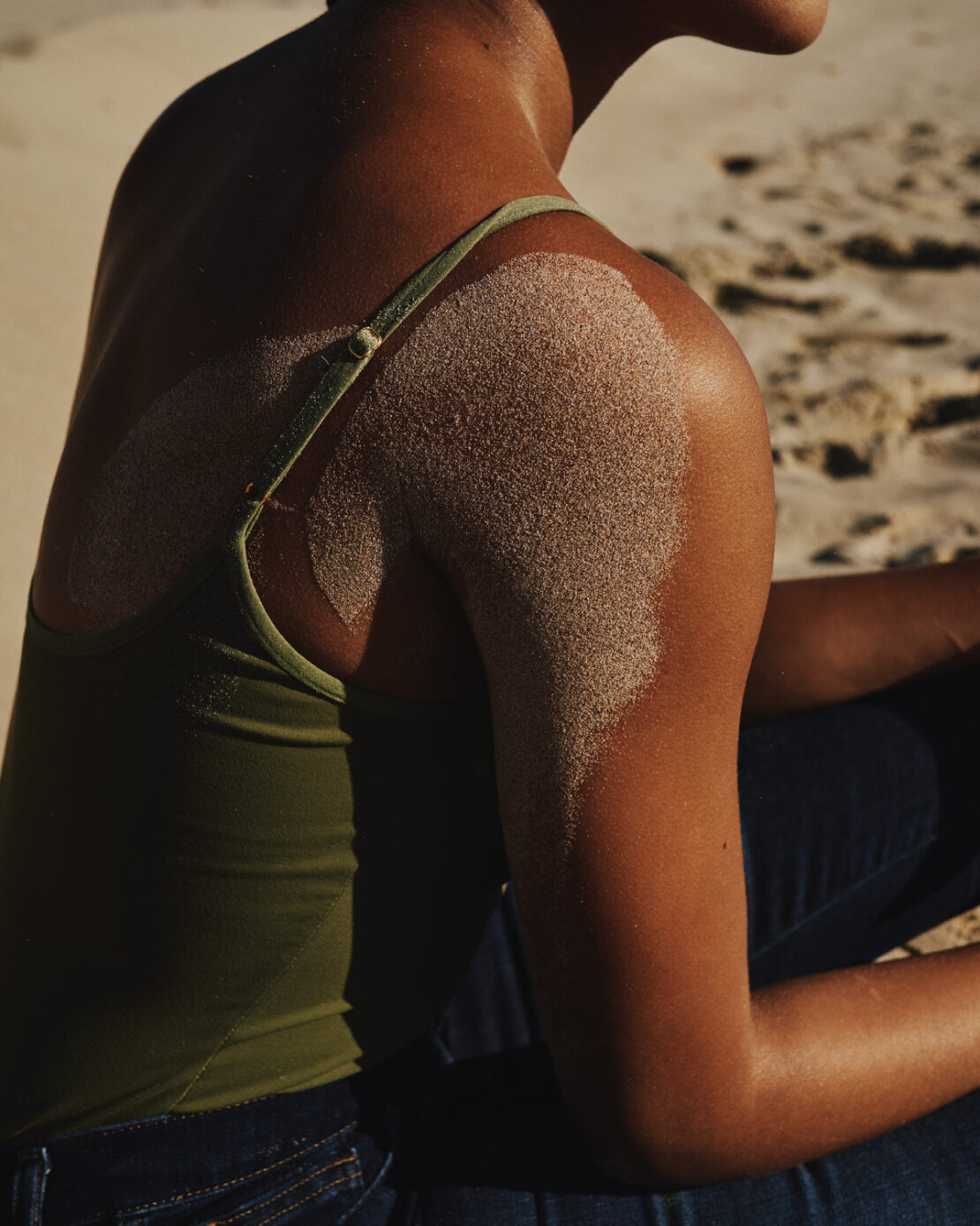 Close-up portrait of a person with sand dusted on their shoulder, wearing an olive green strap top against a sandy beach background.