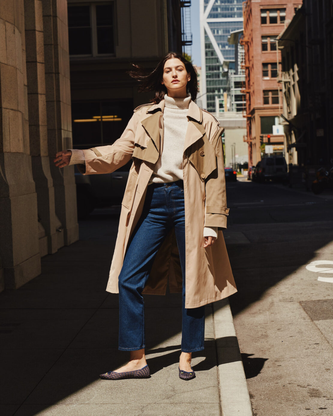 Woman in trench coat walks along city street. She wears jeans and flats. Tall buildings rise in the background on a sunny day.