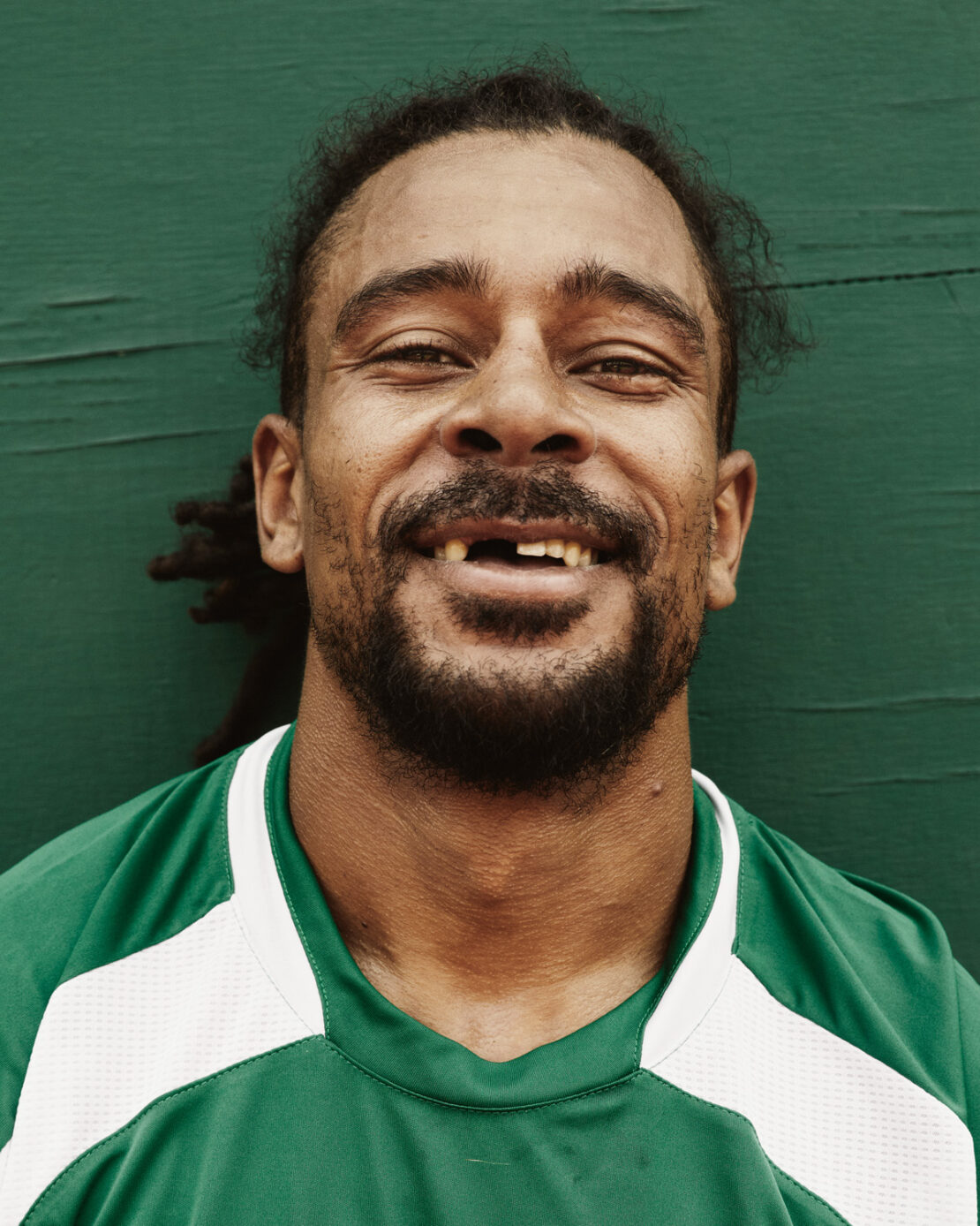 Close-up studio shot of adult male with missing tooth smiling against a green wall, wearing a green and white sport shirt.