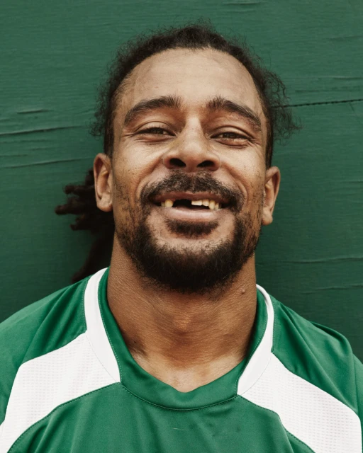 Close-up studio shot of adult male with missing tooth smiling against a green wall, wearing a green and white sport shirt.