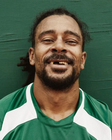 Close-up studio shot of adult male with missing tooth smiling against a green wall, wearing a green and white sport shirt.