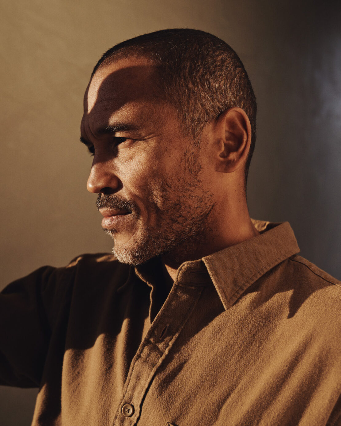 Portrait of a middle-aged man with dark hair and a beard, wearing a tan button-down shirt against a muted background, well-lit.