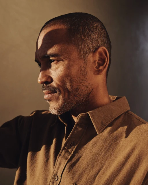 Portrait of a middle-aged man with dark hair and a beard, wearing a tan button-down shirt against a muted background, well-lit.