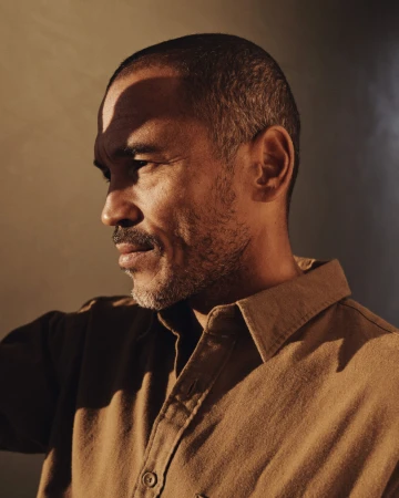 Portrait of a middle-aged man with dark hair and a beard, wearing a tan button-down shirt against a muted background, well-lit.