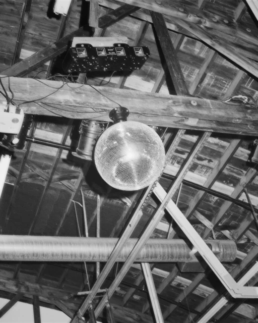 Shiny disco ball hanging in an interior space with exposed rafters and metal ducts. The disco ball is the main focus, creating a vintage ambiance.