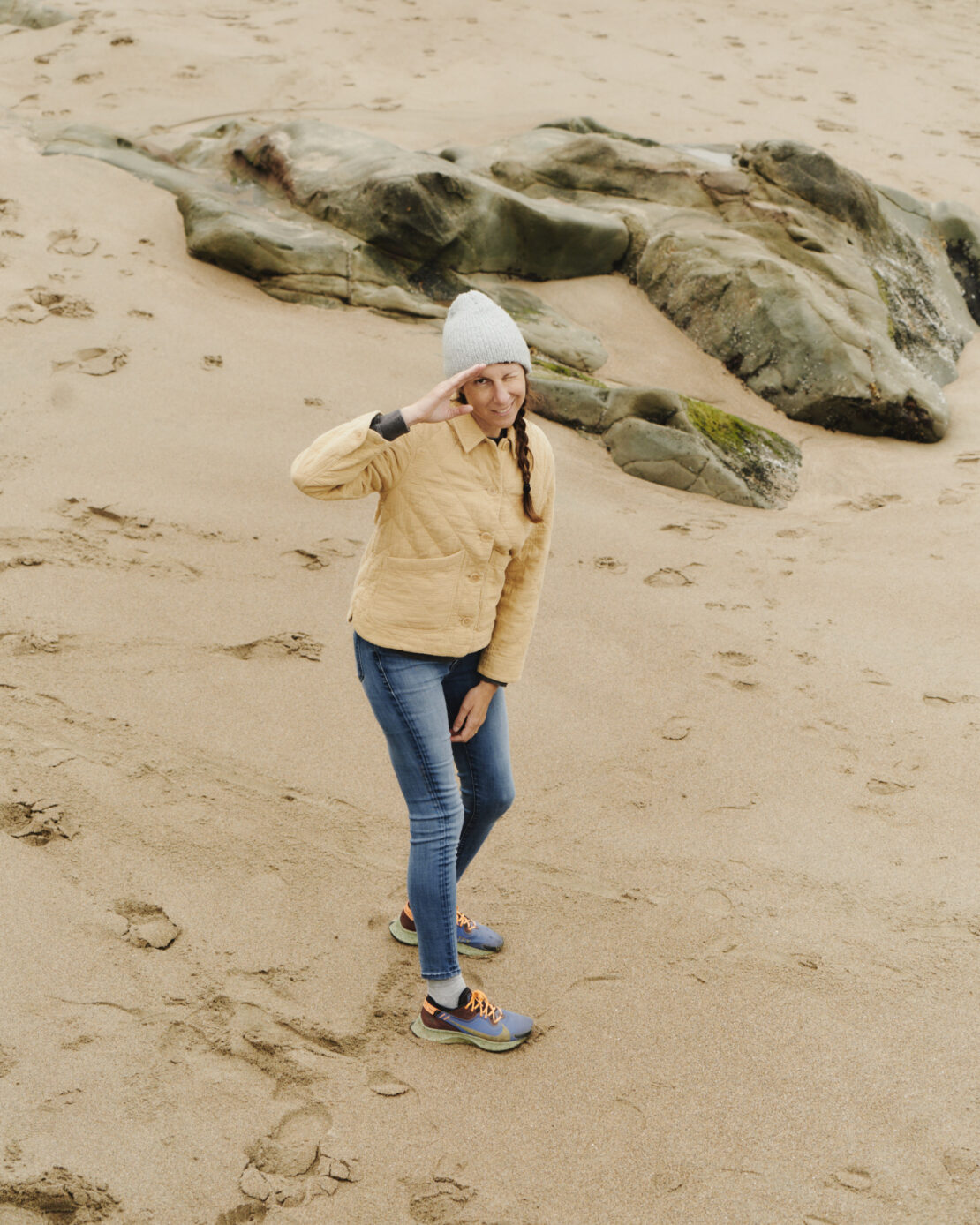 A woman in a yellow jacket and beanie salutes on a sandy beach with rocks in the background, captured in a medium shot at eye-level.