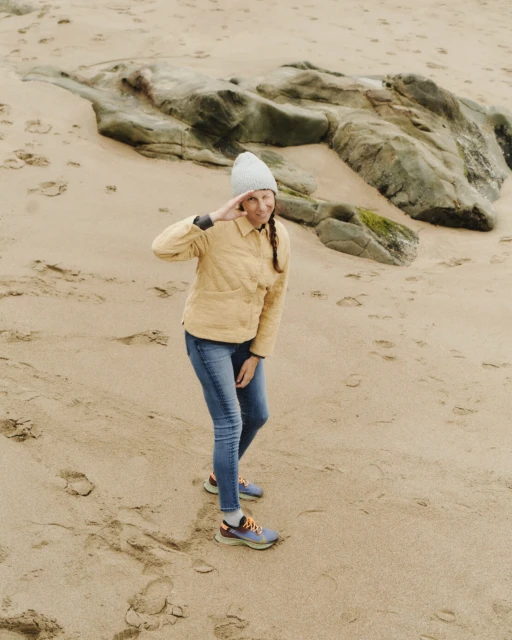 A woman in a yellow jacket and beanie salutes on a sandy beach with rocks in the background, captured in a medium shot at eye-level.
