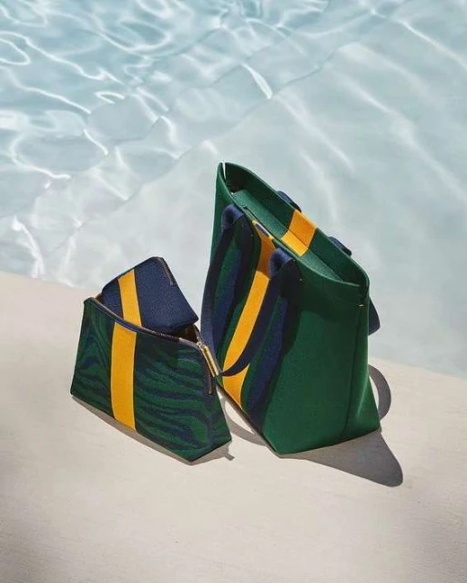 Green and navy striped tote bag and matching makeup bag, featuring a yellow stripe, are set against a sunlit pool background.