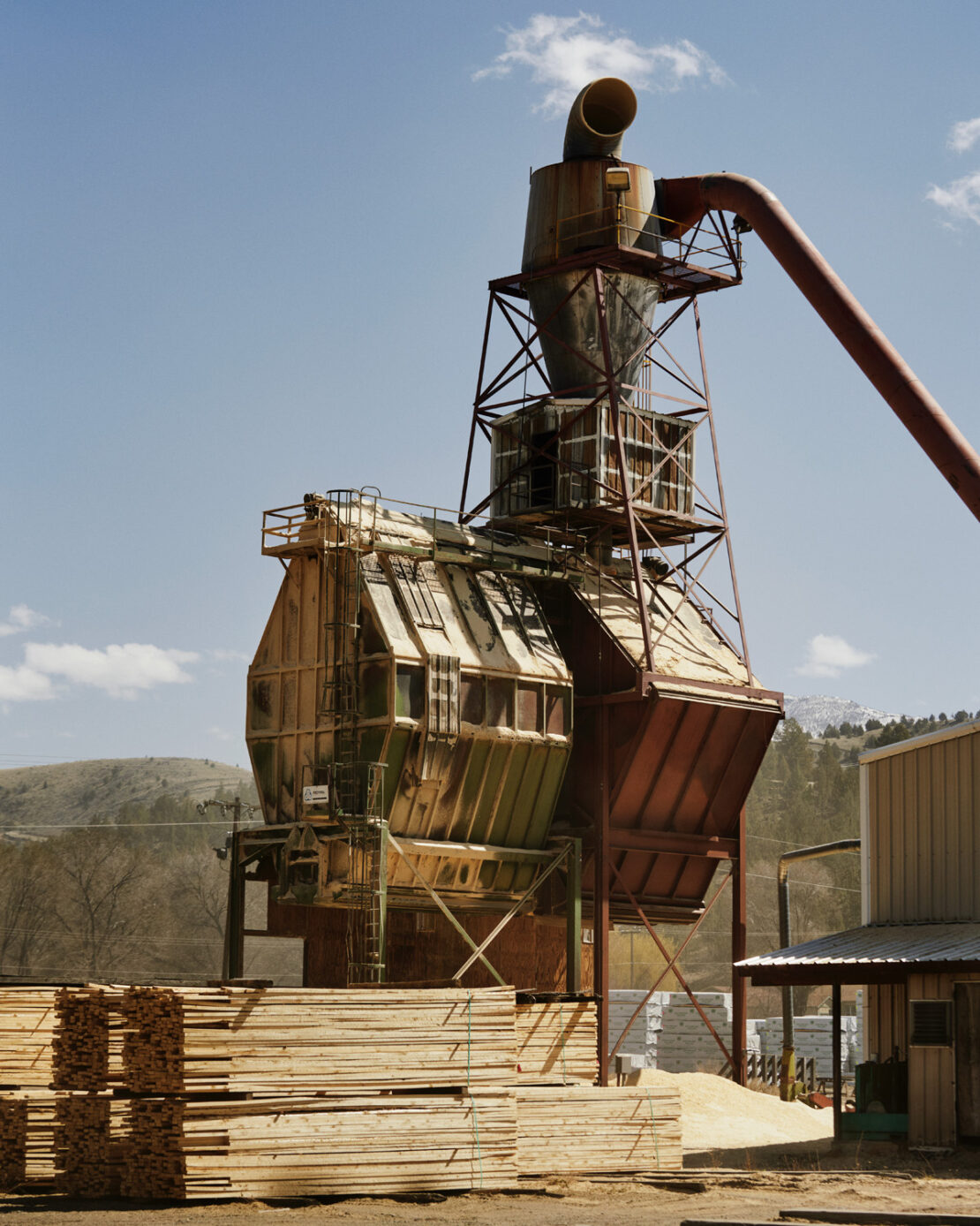 Exterior shot of industrial lumber mill featuring weathered machinery, wooden beams, and a backdrop of trees under a clear sky.