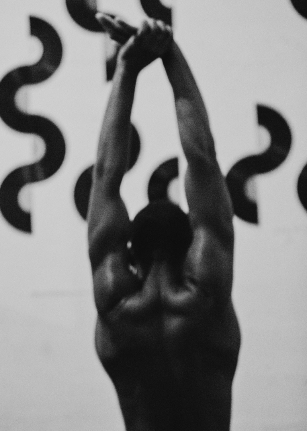 Monochrome shot of a muscular adult male with arms raised overhead, holding a kettlebell in an indoor fitness environment.