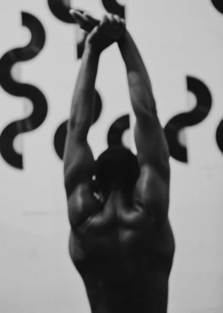 Monochrome shot of a muscular adult male with arms raised overhead, holding a kettlebell in an indoor fitness environment.