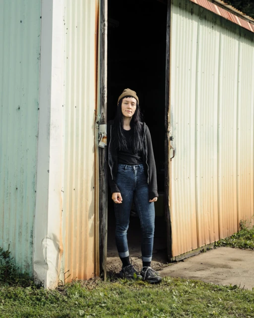 Young woman stands in the doorway of a corrugated metal shed wearing jeans and a beanie in this medium shot with natural light.