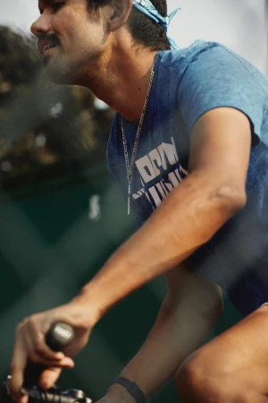 Man wearing a blue t-shirt and bandana riding a bicycle on a sunny day with a green leafy background, captured in a medium shot.