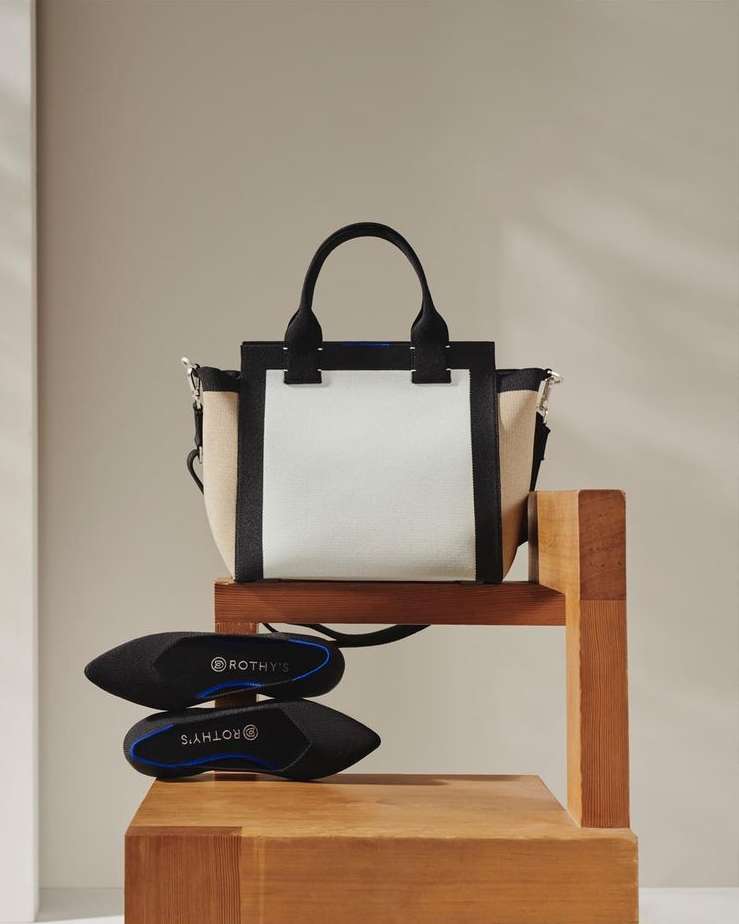 A tan, white, and black Rothy's handbag is staged on a wooden display stand, alongside a pair of black Rothy's pointed-toe flats.