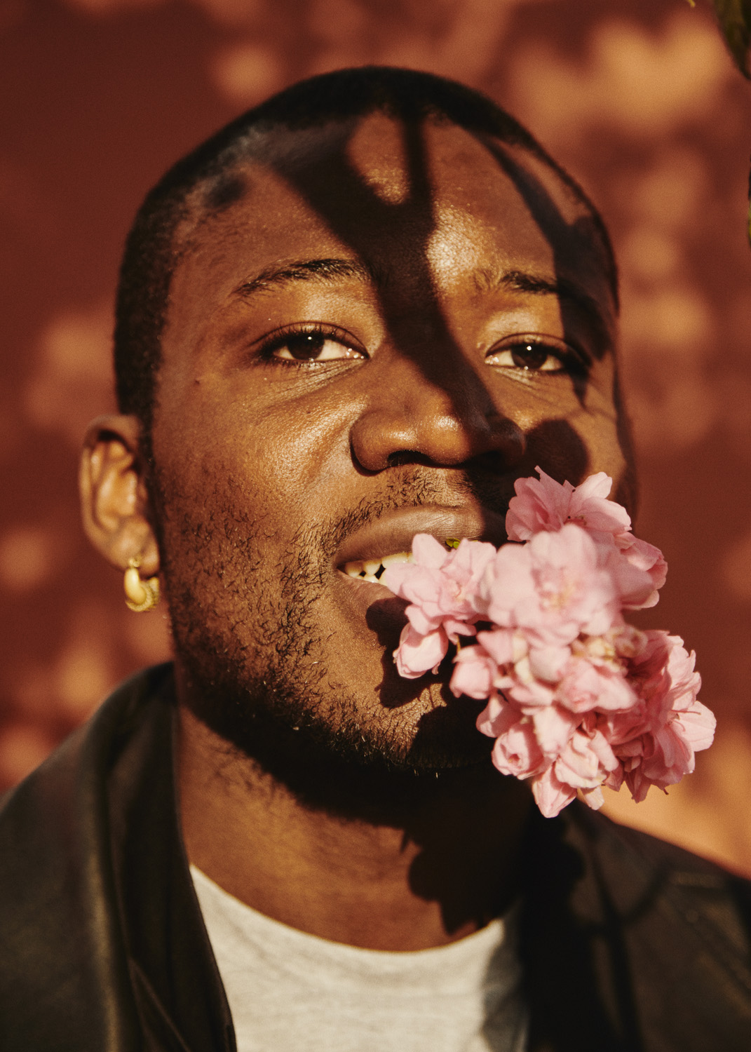 Portrait of a Black man with flowers in his mouth, wearing a black jacket against a textured brown background with tree shadows.