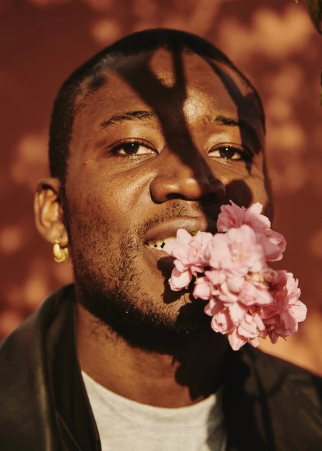 Portrait of a Black man with flowers in his mouth, wearing a black jacket against a textured brown background with tree shadows.