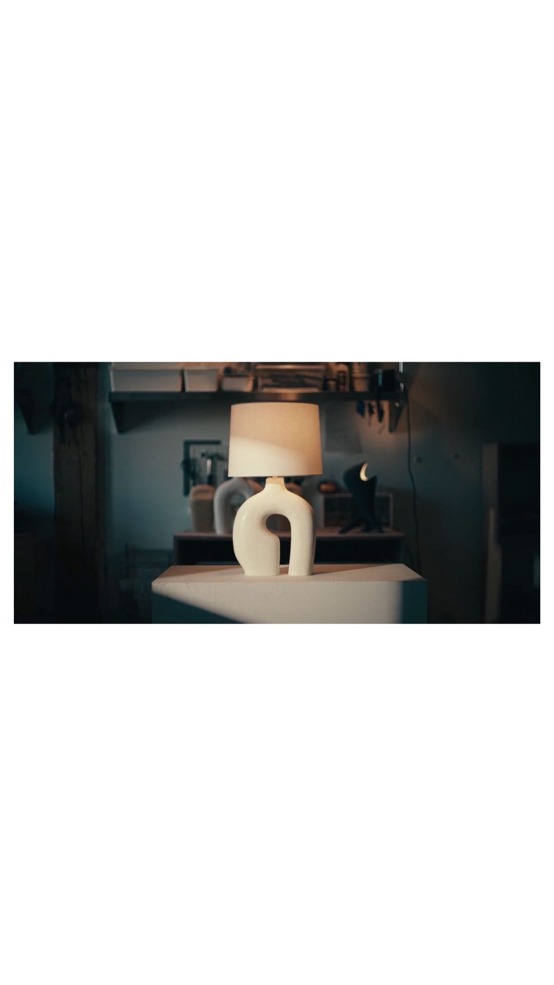 Contemporary table lamp with a white curved base and a lampshade on a table in a dimly lit room showcasing interior design.