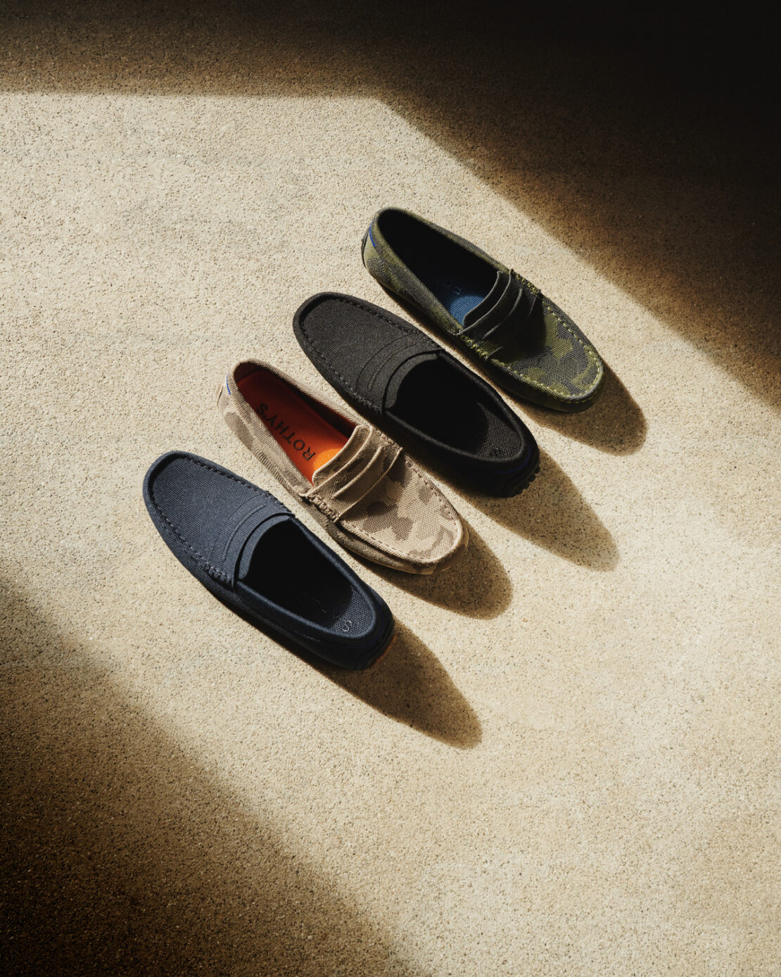 Four Rothy's The Driver loafer-style shoes in navy, beige camouflage, black, and green camo, arranged diagonally on a tan concrete surface.