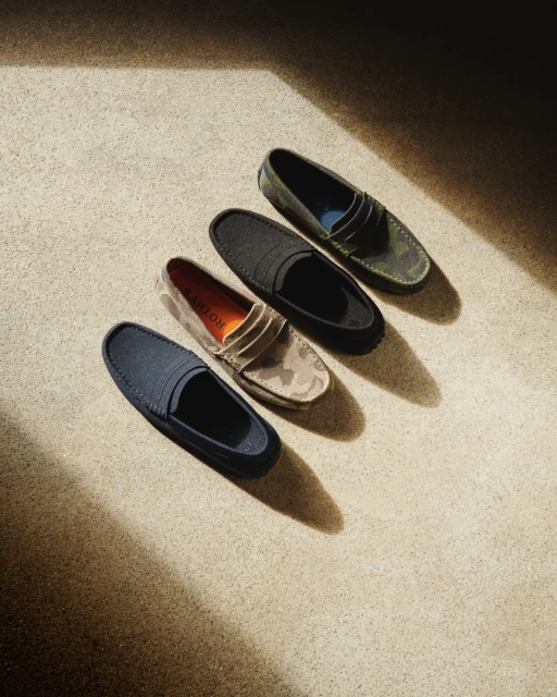 Four Rothy's The Driver loafer-style shoes in navy, beige camouflage, black, and green camo, arranged diagonally on a tan concrete surface.