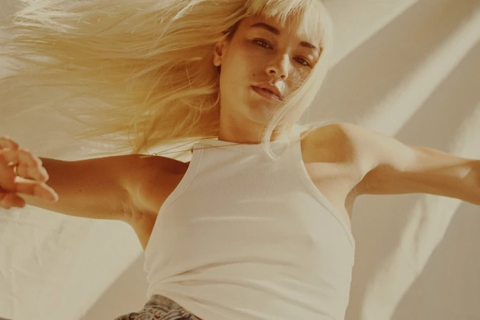 Woman with blonde hair wears white tank top while posing in warm, diffused lighting against a neutral-toned background in a studio setting.