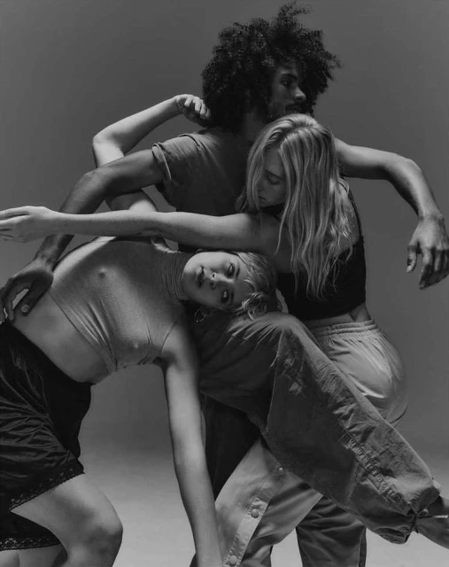 Monochrome image of three adult women and one adult man in a close, dynamic pose, wearing casual clothing, against a neutral grey backdrop.