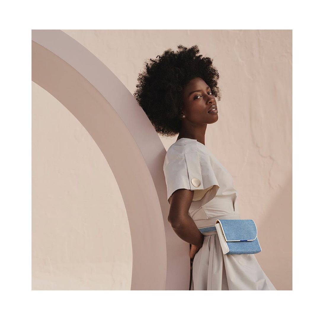 A fashionable Black woman poses in a beige dress with a blue fanny pack in front of a peach wall with an arched doorway for a studio portrait.