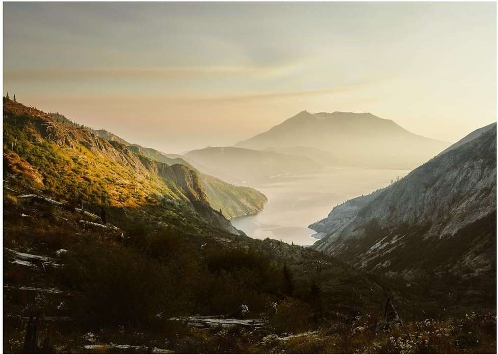 Mountain landscape featuring mist-covered mountains and a valley with a lake, showcasing a scenic outdoor view under soft, hazy light.