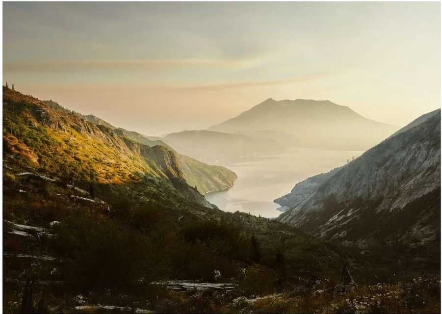 Mountain landscape featuring mist-covered mountains and a valley with a lake, showcasing a scenic outdoor view under soft, hazy light.